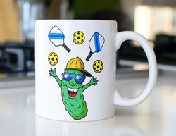 Funny pickleball coffee mug with a pickle caricature juggling paddles and pickleballs