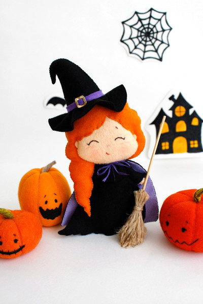 Felt Halloween toy - witch in the pointed hat with a broomstick with orange Halloween pumpkins standing in the background of painted Halloween decorations, fron