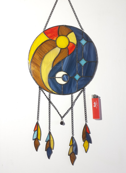 Round stained glass dreamcatcher with blue and yellow Yin Yang symbol is lying on a white background near a red lighter.jpg