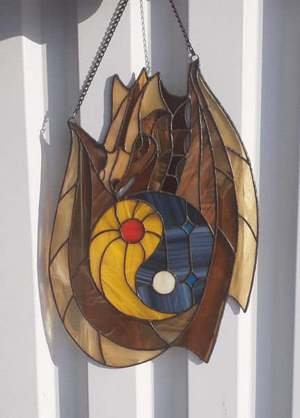Stained glass window panel of a dragon with yin yang symbol in the middle is hanging in front of a white metal fence.jpg