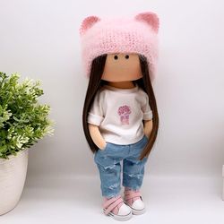 Fashionable rag textile doll in a white T-shirt with a delicate floral print, a pink fluffy hat and ripped jeans