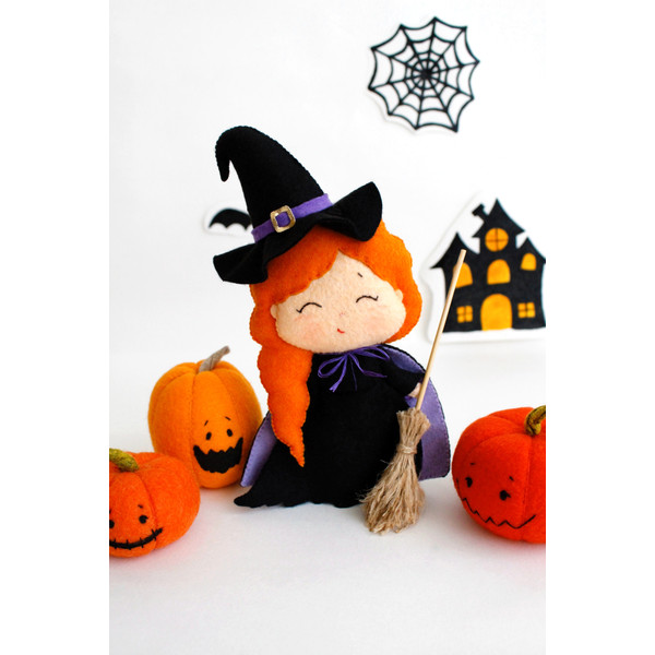 Felt Halloween toy - witch in the pointed hat with a broomstick with orange Halloween pumpkins standing in the background of painted Halloween decorations, fron