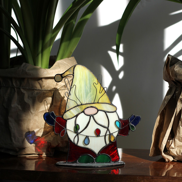 Stained-glass-suncatcher-Christmas-gnome-with-garland-stands-on-a-shelf-next-to-a-flower-pot--The-gnome-has-a-red-glass-coat-blue-glass-mittens-and-a-yellow-gla