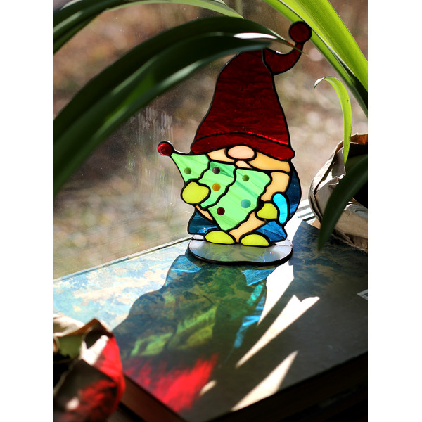 Stained-glass-sun-catcher-Christmas-gnome-with-a-Christmas-tree-in-his-hands-stands-on-a-windowsill-against-the-sunlight-The-sun's-rays-pass-through-the-red-gla