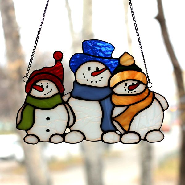 stained-glass-suncatcher-in-the-form-of-three-hugging-snowmen-in-colorful-hats-and-scarves-is-suspended-on-the-window-opposite-the-sun