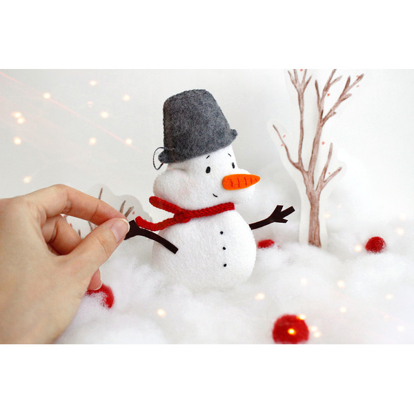 Felt snowman toy with a pail on the head on the background of snow and painted trees