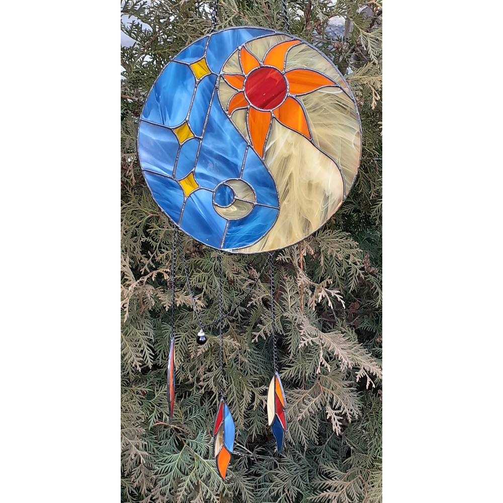 Stained glass Yin Yang dreamcatcher in blue and yellow colors with sun and moon is hanging in front of a Thuja tree.jpg
