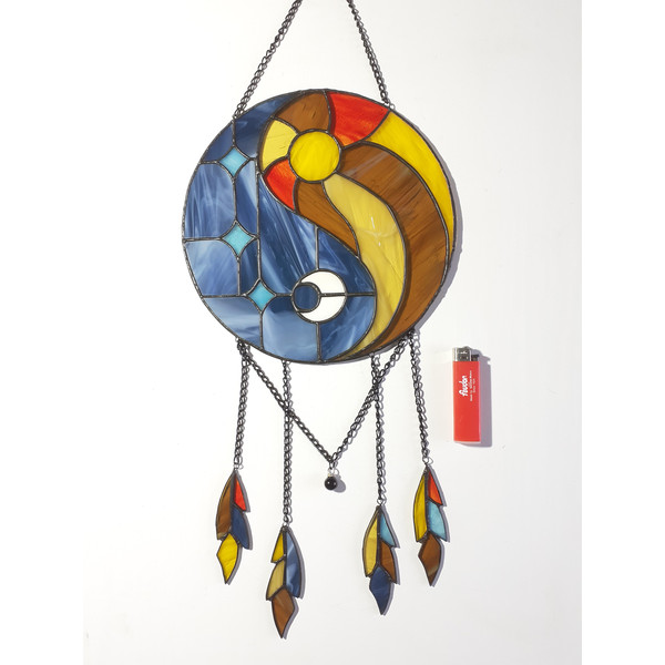 Stained glass dreamcatcher with blue and yellow Yin Yang symbol is lying on a white background near a red lighter.jpg