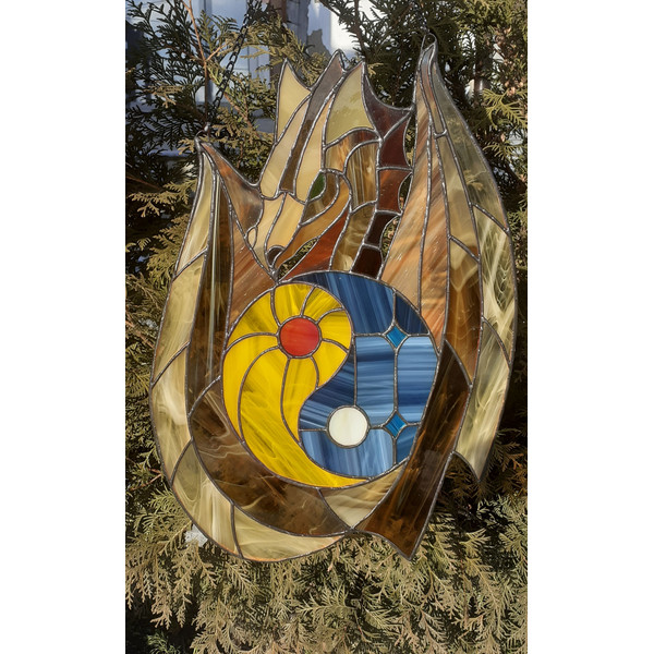 Stained glass window panel of a dragon with yin yang symbol in the middle is hanging in front of a Thuja tree.jpg