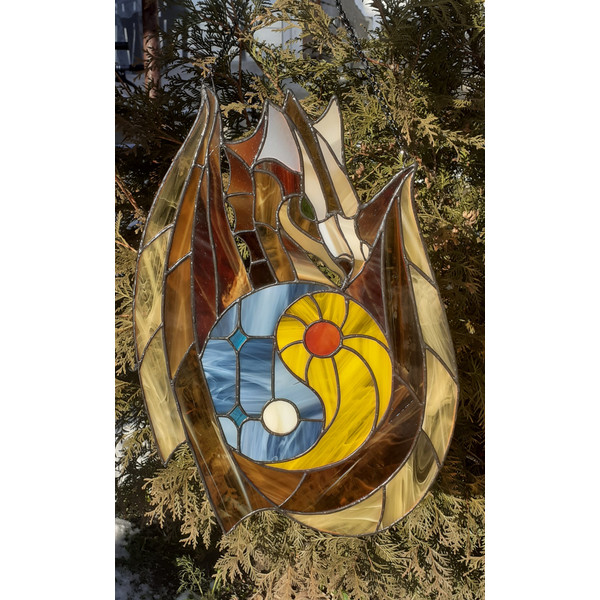 Stained glass window panel of a dragon with sun and moon yin yang in the middle is hanging in front of a Thuja tree.jpg