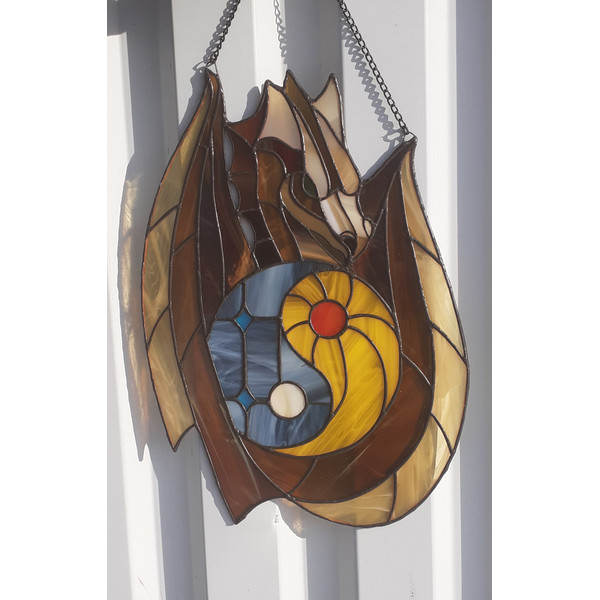 Stained glass window panel of a dragon with sun and moon yin yang in the middle is hanging in front of a white metal fence.jpg