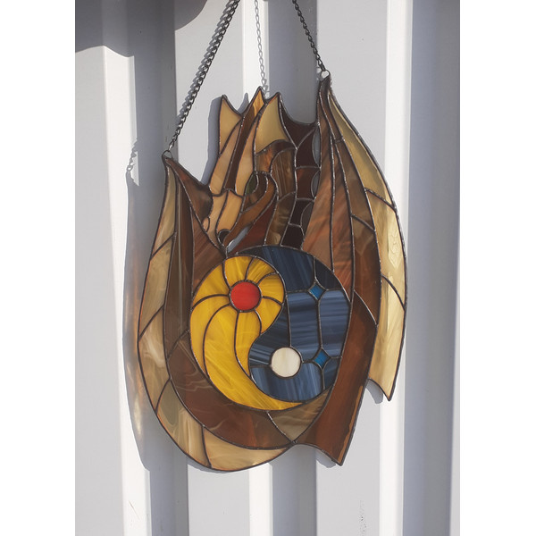 Stained glass window panel of a dragon with yin yang symbol in the middle is hanging in front of a white metal fence.jpg