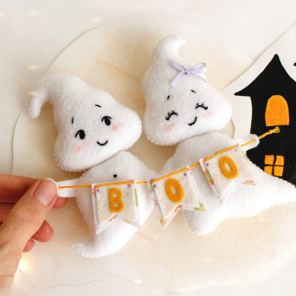 Felt ghost boy and girl with BOO garland laying in the author's hand against the background of painted Halloween decorations, top view