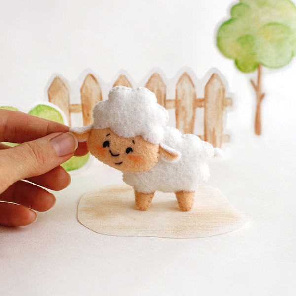 Felt farm animal - cute sheep in the authors hand stands in the background of painted tree and fence