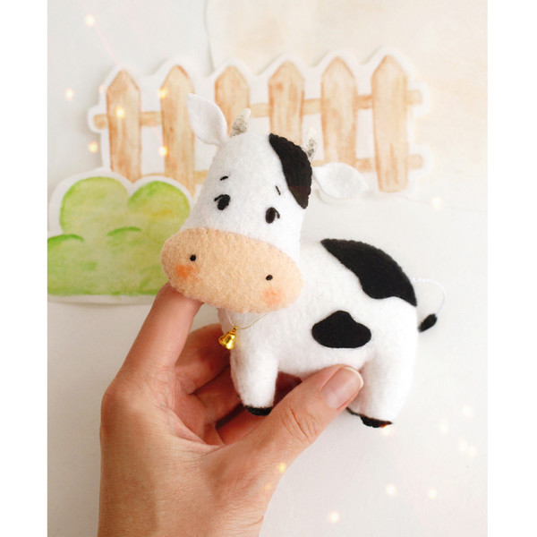 Felt farm animal - cute white and black cow in the authors hand in the background of painted tree and fence